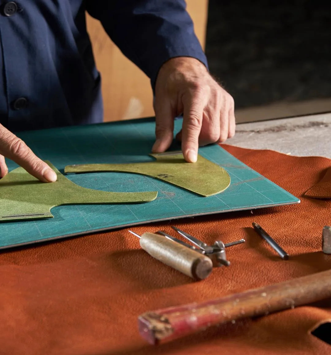 Le mani di un artigiano che tagliano a mano una tomaia in pelle verde su un pellame color cuoio, con lesina e compasso posati accanto